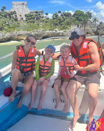 Family wearing bright orange life jackets smiling on a small boat off a tropical rocky coastline with palm trees and ancient cliffside stone ruins under a bright blue sky