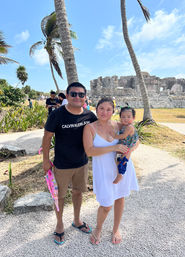 Sunny family portrait at Tulum, Mexico: parents and a toddler posing by palm trees and coastal Mayan ruins under a bright blue sky