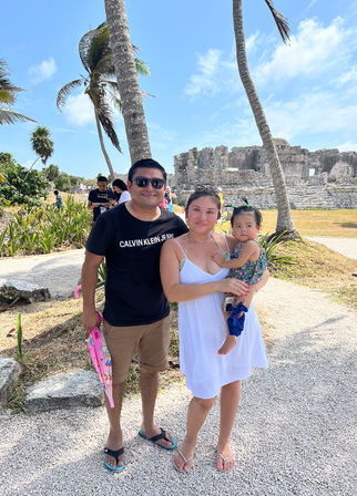 Sunny family portrait at Tulum, Mexico: parents and a toddler posing by palm trees and coastal Mayan ruins under a bright blue sky