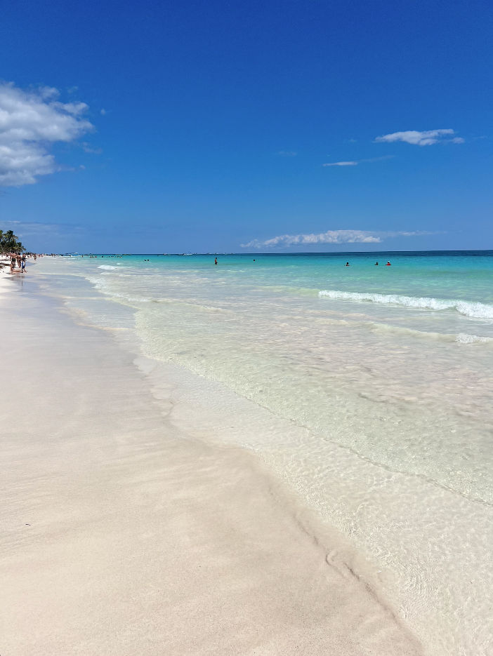 Sunlit tropical beach with powder-white sand and clear turquoise water, gentle waves lapping the shore under a bright blue sky, a few distant swimmers and palm trees along the coastline.
