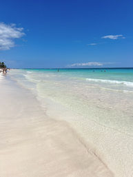 Sunlit tropical beach with powder-white sand and clear turquoise water, gentle waves lapping the shore under a bright blue sky, a few distant swimmers and palm trees along the coastline.