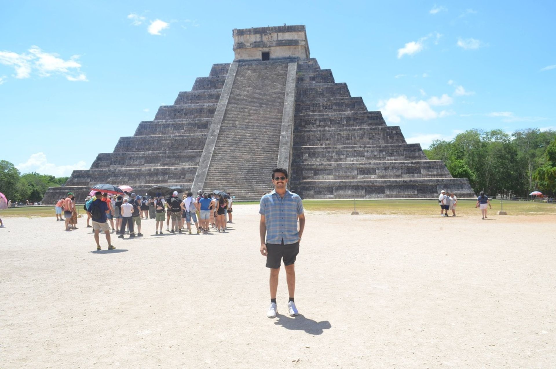 Person posing in front of the El Castillo pyramid at Chichén Itzá, Yucatán, Mexico, with tourists, bright sun and a clear blue sky.