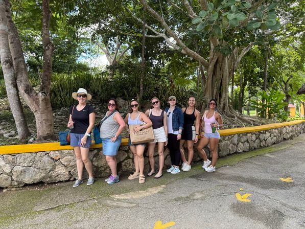 Eight women in casual summer outfits posing along a low stone wall under a large tree with exposed roots in a lush tropical park, smiling on a shaded paved walkway during a summer outing.