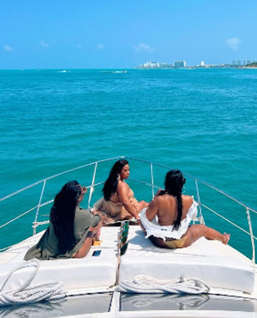 Three women lounging on a yacht bow, soaking up the sun over turquoise ocean with a distant coastal skyline under a bright blue sky.