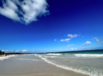 White-sand tropical beach with gentle turquoise waves lapping the shore, clear deep-blue sky dotted with fluffy white clouds and small groups of people in the distance — sunny seaside scene.