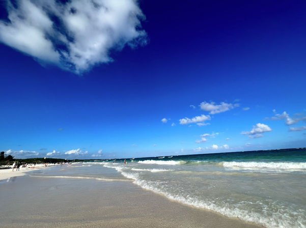 White-sand tropical beach with gentle turquoise waves lapping the shore, clear deep-blue sky dotted with fluffy white clouds and small groups of people in the distance — sunny seaside scene.