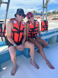 Two smiling people in orange life jackets and sunglasses sit barefoot on a blue-and-white tour boat off a sunny tropical beach with palm trees and thatched umbrellas, vacation vibe.