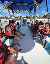 Smiling kids and adults in bright orange life vests seated on a shaded excursion boat in clear turquoise water approaching a palm‑lined tropical beach under a sunny blue sky.