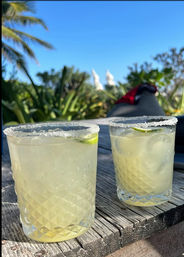 Two salt-rimmed margarita glasses with lime wedges on a weathered wooden table, palm trees and clear blue sky in a sunny tropical beachside outdoor setting.