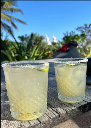 Two salt-rimmed margarita glasses with lime wedges on a weathered wooden table, palm trees and clear blue sky in a sunny tropical beachside outdoor setting.