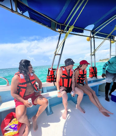 Three passengers in bright orange life jackets chill under a blue canopy on a day boat, sipping drinks as turquoise tropical water and a sandy island pass by.