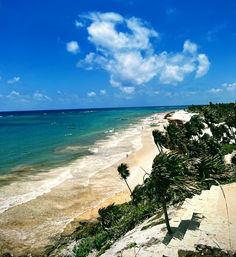 Aerial view of a tropical coastline with turquoise waters, white-sand beach dotted with small boats, swaying palm trees on rocky cliffs and stone steps under a bright blue sky with fluffy clouds