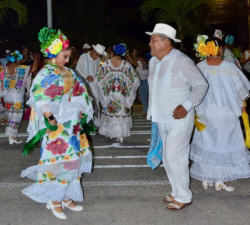 Night street festival in Mexico with dancers: woman in colorful embroidered white huipil and floral headpiece twirling, man in white guayabera and hat dancing, other lace-trimmed traditional costumes in background.