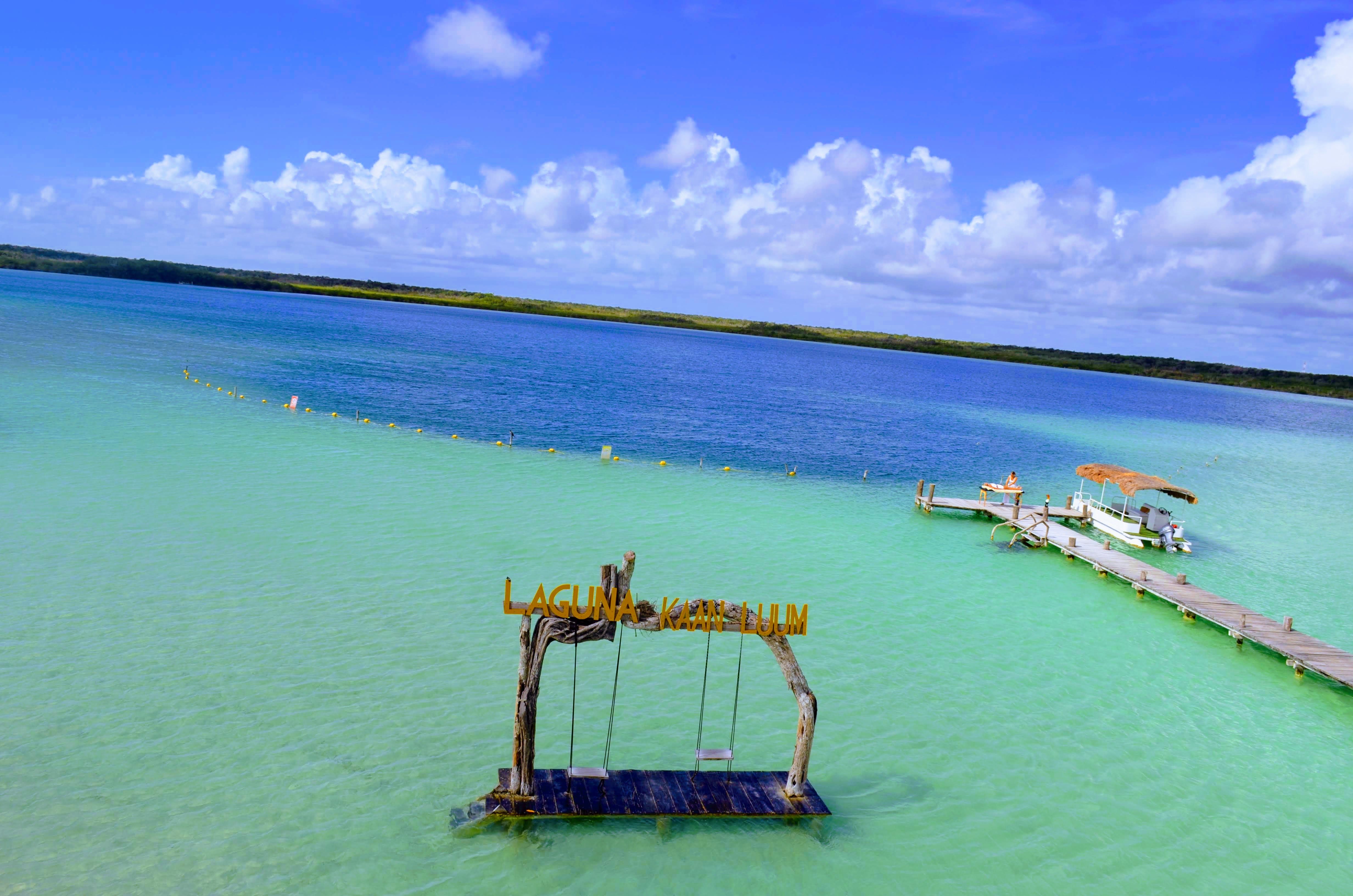 Aerial view of a tropical turquoise lagoon with a rustic wooden swing platform in shallow water, a long wooden dock with a thatched-roof boat, and fluffy white clouds under a bright blue sky.