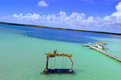 Aerial view of a tropical turquoise lagoon with a rustic wooden swing platform in shallow water, a long wooden dock with a thatched-roof boat, and fluffy white clouds under a bright blue sky.