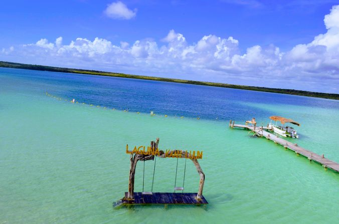 Aerial view of a tropical turquoise lagoon with a rustic wooden swing platform in shallow water, a long wooden dock with a thatched-roof boat, and fluffy white clouds under a bright blue sky.