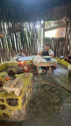 Person making handmade tortillas over a wood-fired comal inside a rustic palapa-style kitchen with clay pots, wooden table, mortar and slatted wooden walls.