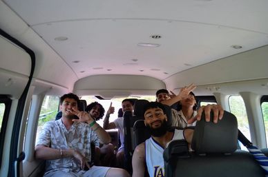 Six young adults smiling and waving inside a passenger van on a daytime road trip, casual summer clothes, thumbs-up and playful poses, interior view with green countryside visible through windows.