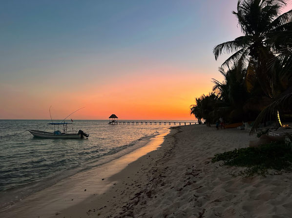 Serene tropical sunset over a sandy beach with a small fishing boat in calm water, a long wooden pier with a thatched gazebo, and palm trees silhouetted along the shoreline.