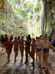 Group of women in swimsuits posing on a wet stone ledge inside a sunlit tropical cenote sinkhole, surrounded by long hanging vines and an emerald-blue pool.