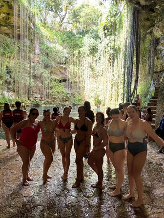 Group of women in swimsuits posing on a wet stone ledge inside a sunlit tropical cenote sinkhole, surrounded by long hanging vines and an emerald-blue pool.