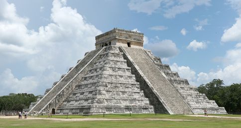 El Castillo stepped stone pyramid at Chichén Itzá with central staircase, grassy plaza and small groups of visitors under a blue sky — Yucatán, Mexico