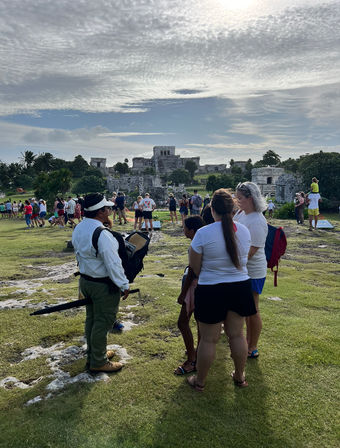 Tourists and a guide on a grassy overlook facing the ancient Mayan stone ruins of Tulum, Mexico, under a dramatic cloud-filled sky.