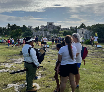Tourists and a guide on a grassy overlook facing the ancient Mayan stone ruins of Tulum, Mexico, under a dramatic cloud-filled sky.