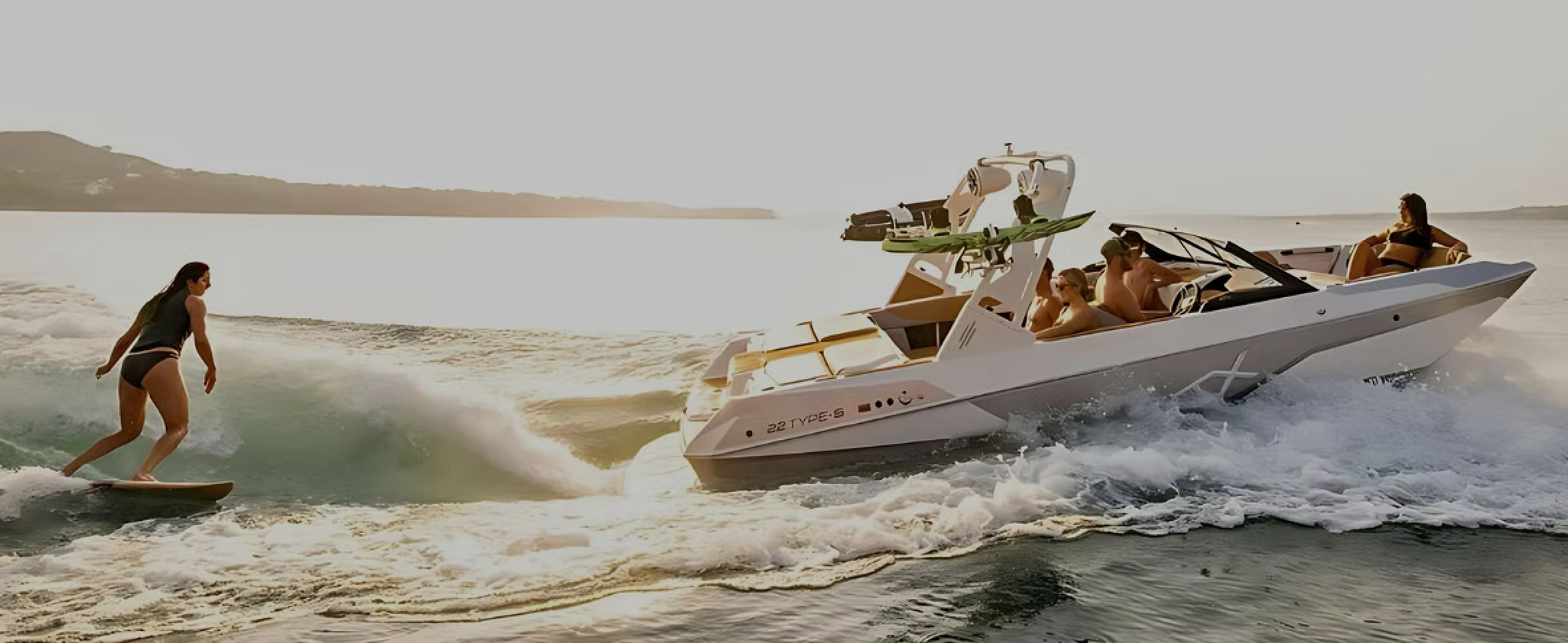 Wakesurfer carving a wake behind a white speedboat as passengers relax, cruising across sunlit coastal waters at golden hour.