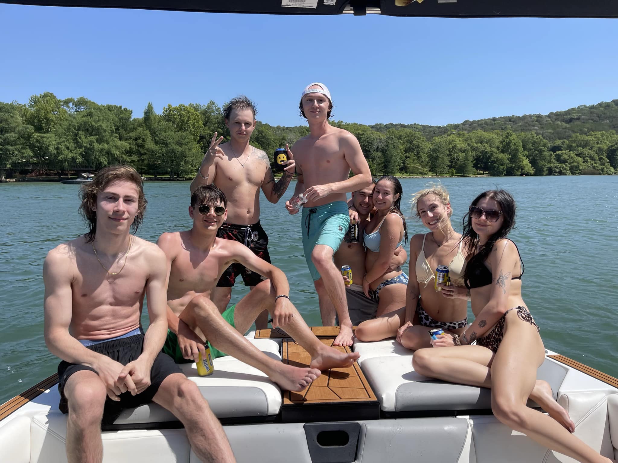 Group of eight friends in swimwear posing with drinks on the stern of a boat on a sunny summer lake, tree-lined shoreline and rolling hills in the background.