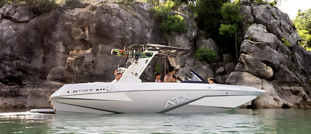 White wakeboard-style motorboat with a tower and board on top carrying a group of people in a secluded rocky cove, anchored on calm green lake water under sunlit trees.
