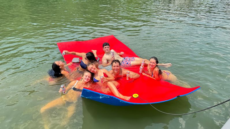 Seven friends lounging and smiling on a bright red floating mat in green lake water, holding canned drinks and enjoying a sunny summer day while tethered to a boat by a rope.