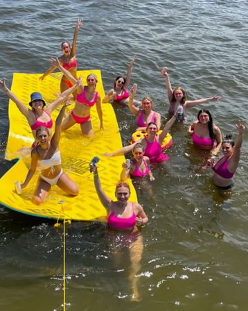 Group of women in bright pink swimsuits enjoying a sunny lake party on a large yellow floating mat, waving and holding drinks