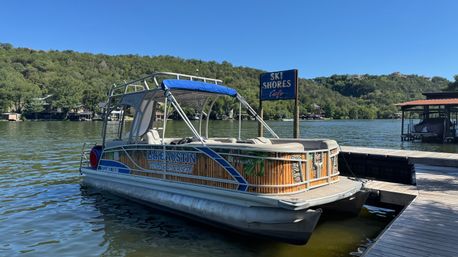 Pontoon boat with blue canopy moored at a wooden dock on a sunny lake, tree‑covered hills and clear blue sky in the background.