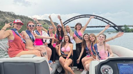 Cheerful women in swimsuits and sashes partying on a motorboat with arms raised, cruising Lake Austin beneath the Pennybacker (360) Bridge in Austin, Texas.