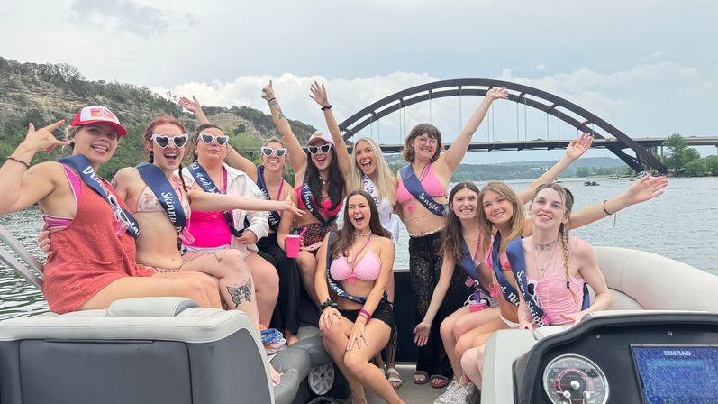 Cheerful women in swimsuits and sashes partying on a motorboat with arms raised, cruising Lake Austin beneath the Pennybacker (360) Bridge in Austin, Texas.