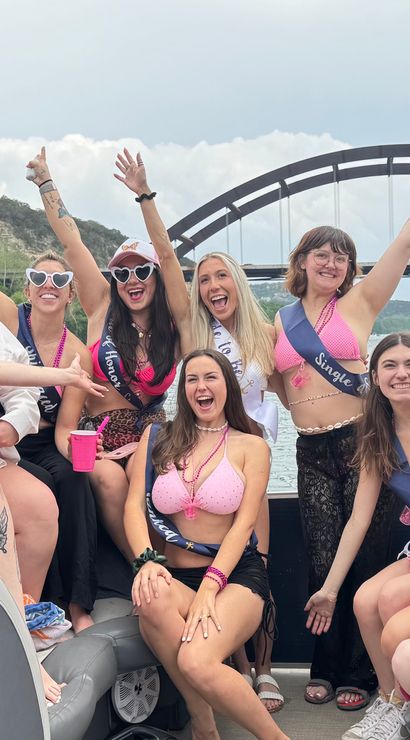 Cheerful women in swimsuits and sashes partying on a motorboat with arms raised, cruising Lake Austin beneath the Pennybacker (360) Bridge in Austin, Texas.
