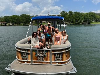 Group of friends relaxing and smiling on a tiki-decor pontoon boat with a blue canopy, wearing swimsuits and cover-ups on a sunny lake with tree-lined shoreline and waterfront homes.