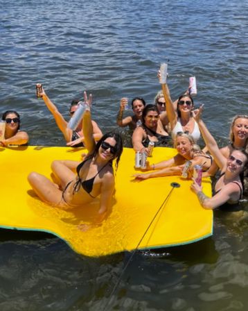 Group of women in swimsuits on a bright yellow floating pad in a lake, holding canned drinks and cheering during a sunny summer lake party