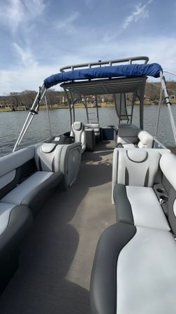 Pontoon boat interior with gray and white cushioned seating and blue bimini top, docked on a calm lake near a lakeside neighborhood under a partly cloudy sky.