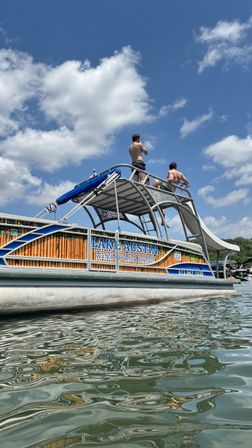 Pontoon boat with upper-deck waterslide and two shirtless people standing on top under a bright blue sky, reflected in rippling lake water.