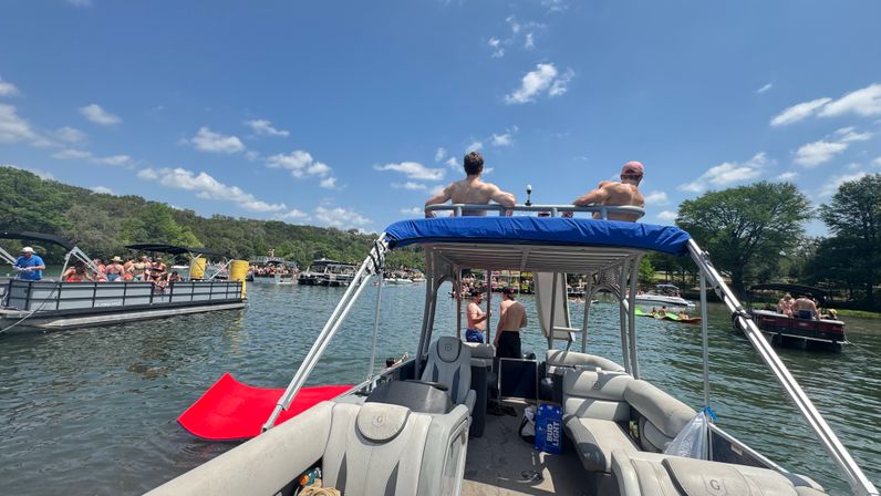 Sunny summer lake scene from a pontoon boat: people on nearby pontoons, a red water slide, blue sky and tree-lined shore.