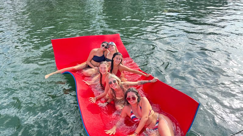 Six women in bikinis lounging and smiling on a bright red floating foam mat on a lake, splashing and enjoying a sunny summer day