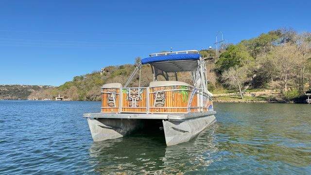 Tiki-decor pontoon boat with blue canopy floating on a calm lake near a tree-covered hillside under a clear blue sky
