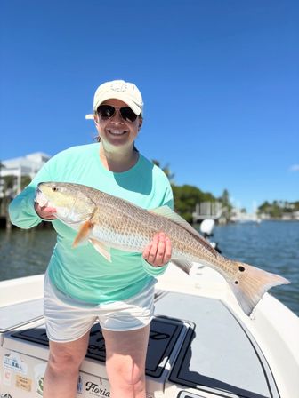 Smiling angler in a cap and sunglasses holding a large redfish (red drum) with tail spot on a boat in Florida backwaters under a bright blue sky