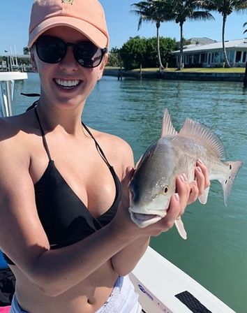 Smiling angler in a cap and sunglasses holding a fresh catch on a boat in a sunny tropical waterfront canal with palm trees and waterfront homes