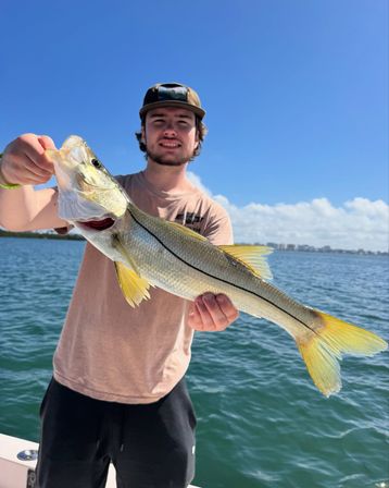 Young angler holding a yellow‑tailed snook on a sunny boat trip, with blue coastal waters and a clear sky in the background