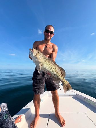 Angler on a small boat holding a large mottled grouper — big catch during offshore saltwater fishing on a calm blue sea under a clear sunny sky