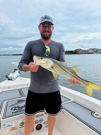 Angler on a boat holding a freshly caught snook with a yellow tail, calm inshore waters and waterfront homes under a cloudy sky — Florida inshore snook fishing.