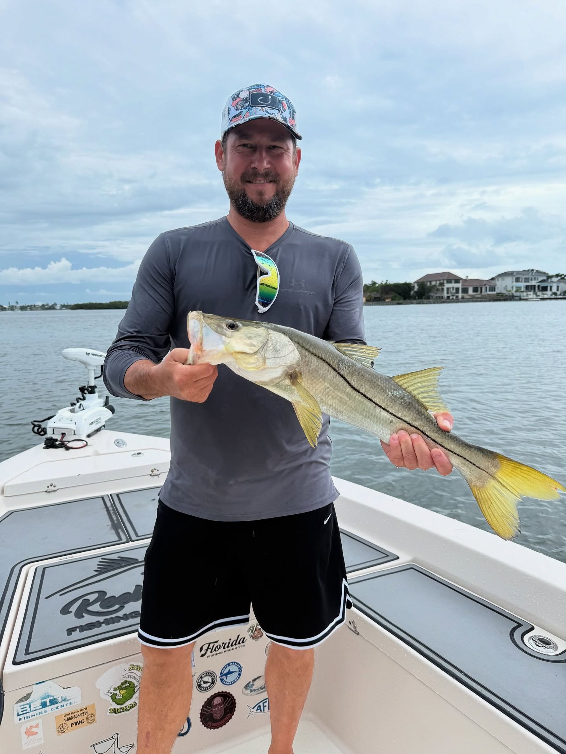 Angler on a boat holding a freshly caught snook with a yellow tail, calm inshore waters and waterfront homes under a cloudy sky — Florida inshore snook fishing.